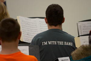 Photo of a student from the back. He is sitting and looking at sheet music. The back of his t-shirt reads, “I’m with the band.”