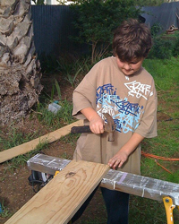 A photograph of a boy hammering a nail into a board on a workhorse, outdoors.