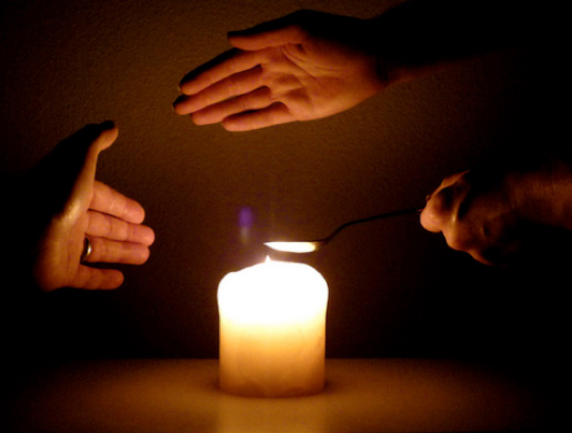 candle surrounded by three hands. One above it, one to the left side and one holding a metal spoon in the flame