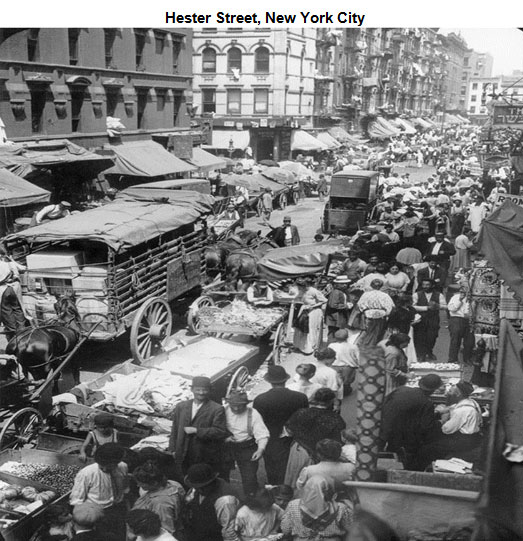 Image of Hester Street in New York City. The street is crowded on both sides with throngs of people on each side. There are vendors that line each side and horse carriages are riding through the middle. 