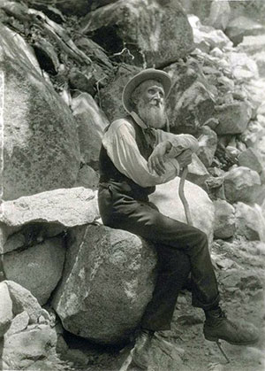A photograph of naturalist John Muir. In this photo he is outdoors seated on a rock, leaning on a cane. He is in his later years with a full beard wearing a hat.