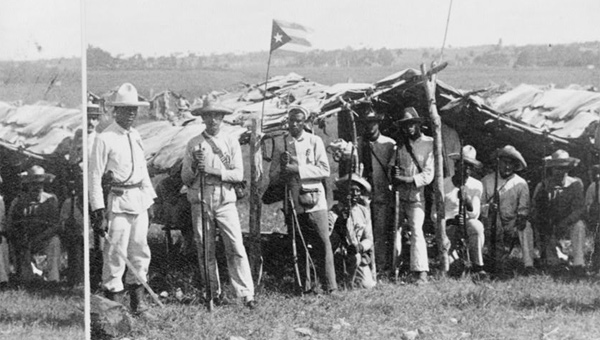 This is a photo of Cuban soldiers posing in front of a makeshift camp with a Cuban flag flying above. Most are dressed regular white shirts and pants. Some are armed with weapons, including a strand of bullets draped across their bodies.