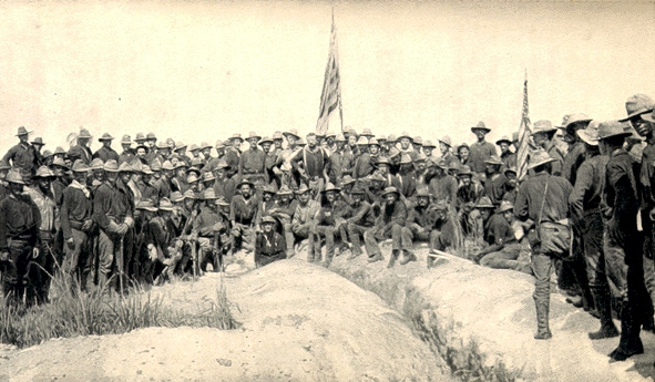 Photo of the Rough Riders, 1st Volunteer Cavalry. Theodore Roosevelt can be seen in the middle of the photo with an American flag raised several rows behind him. The men are pictured on top of the 'San Juan Hill'.