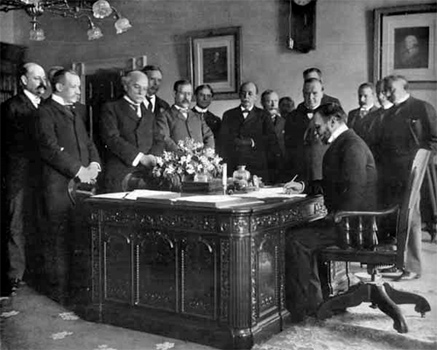 Photo of Secretary of state John Hay signing the Treaty of Paris. He is seated at a desk while several other men are standing around the desk watching him.