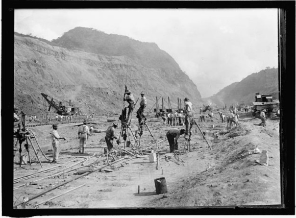 Image of men working at the bottom of the canal. Some of the men are standing on a mechanism that is dug deeply into the ground. There is a crane on the left side of the photo and a crudely constructed one and a half story building exists in the background on the right.