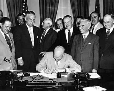 Image of President Eisenhower signing the bill. There are several men standing behind him, watching him sign. 