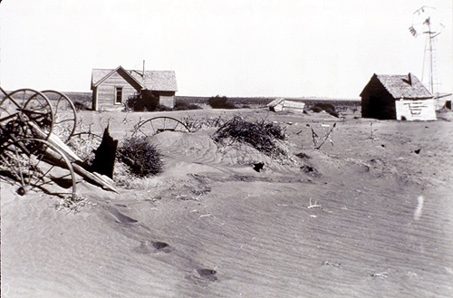 Photo of an abandoned farm that has been covered in dirt. The tractors in the forefront of the photo are almost completely covered by the dust. There is a house in the background and a barn in the back. A windmill can be seen far in the background.