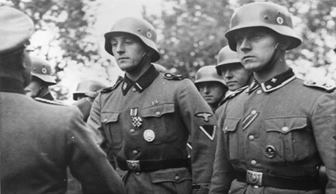 A photograph of German soldiers being decorated. These soldiers are wearing SS uniforms with SS collar patches, belt buckles and helmets with swastika insignia.