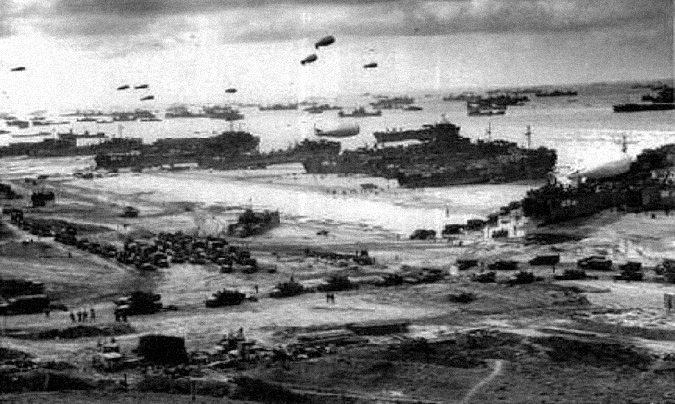 Image of a panoramic view of the D-day Invasion; the view is of the beach of Normandy, France. There are carrier ships lined up on the beach and at least two dozen battleships and carrier ships are positioned offshore. There are rows of tanks and trucks on the ground and men can be seen in a various locations on the ground. There are also aircrafts in the air.