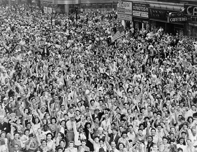 Image of a bird's eye view photo of a street intersection that is crowded with hundreds of people who are cheering; some are holding American flags.