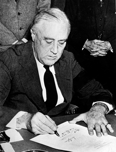 A photograph of US President Franklin D. Roosevelt signing the Declaration of War against Japan in 1941. He is man in late middle age wearing a suit, seated at a table surrounded by other men.