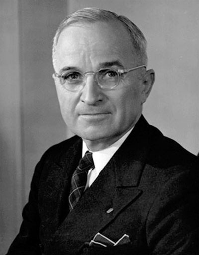 A photograph portrait of President Harry Truman. He is man in late middle age wearing glasses, a suit and tie.