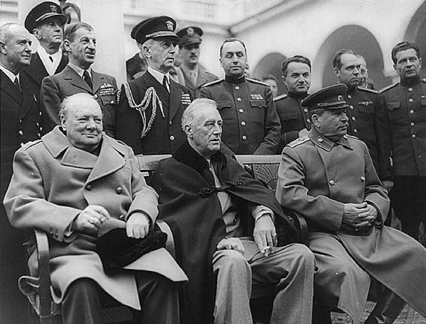 Image of the Big Three seated on a bench while several men representing militaries of different countries are standing behind the bench. Seated on the bench from left to right are: Winston Churchill, Franklin D. Roosevelt, and Josef Stalin.