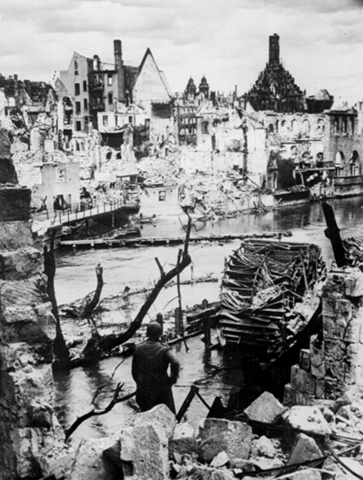 Image of a photo of the German city of Nuremberg on April 20, 1945. The image is of a city with building destroyed by war. There is a man sitting on the bank of the water with a destroyed boat in front of him.