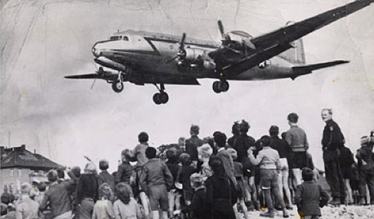 Image of a US Air Force C-47 airplane, flying low over a large crowd of people.