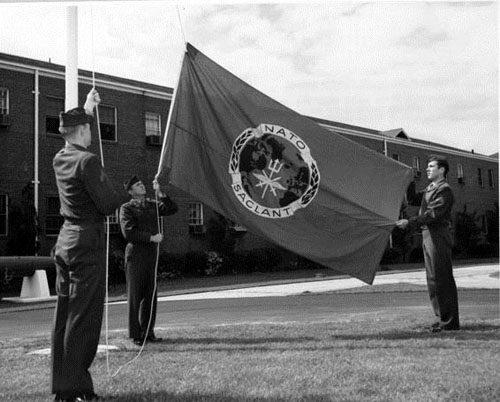 A photograph of three NATO soldiers raising the NATO flag outside of a barracks style building.
