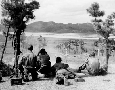 Photo of four soldiers on top of a hill overlooking a river bank (three are sitting), one soldier (lying) is firing on distant Korean patrols with a machine gun.