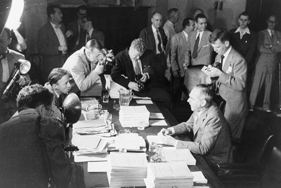 Photo of U.S. Secretary of State Dean Acheson who is seated at a table covered with bound reports. He is surrounded by reporters and cameramen.