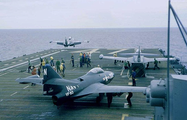 Photo of the back of an aircraft carrier in the middle of a body of water. There are three visible U.S. naval aircrafts, one is taking flight. There are several servicemen on the deck.