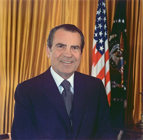 A photograph of US President Richard Nixon standing in front of an American flag. He is a man in late middle age wearing a suit and tie.