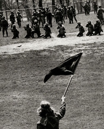 A photograph of war protesters at Kent State University in Ohio. They are waving signs and carrying flags in front of US soldiers that are aiming rifles at them.