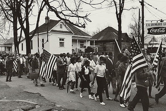 Image of dozens of men and women walking along a street, some are carrying American flags.