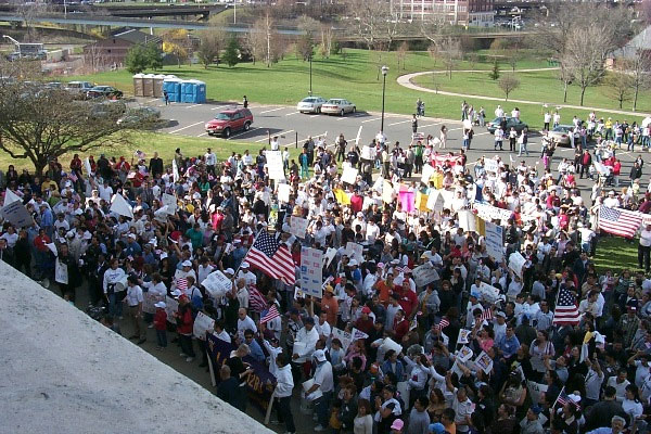 Image of hundreds of people standing on the lawn of a building. Some are carrying signs and other carrying American flags.