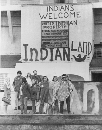  Image of several men and women standing outside Alcatraz with a sign in the background welcoming all Indians