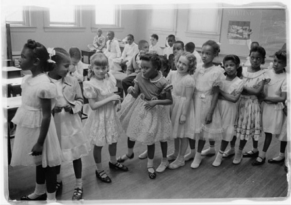Image of a line of grade school aged girls (both black and white) standing in a line in a classroom. The boys are seated at desks in the background.