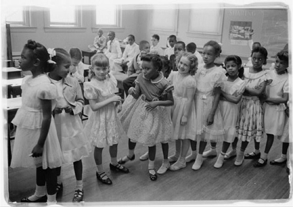 Image of a line of grade school aged girls (both black and white) standing in a line in a classroom. The boys are seated at desks in the background.