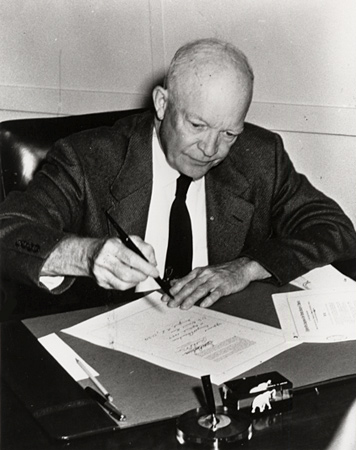 Image of Dwight Eisenhower signing the civil rights act of 1957 as he sits at his desk. 