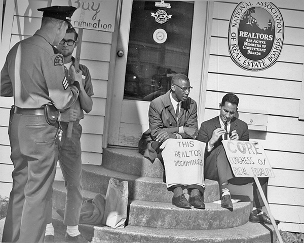 Image of two men holding signs (one reads: 'This realtor discriminates' and the other 'CORE Congress of racial equality') as they sit on the steps of a real estate office. A police officer is speaking to another man who is holding a sign. 