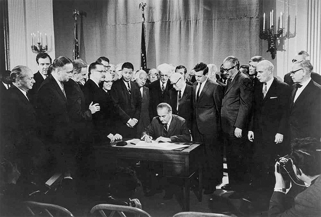Image of President LBJ signing the Civil rights Act of 1968 at his desk. He is surrounded by several men assembled in three rows, watching him sign the document. There are cameramen kneeling in front, taking picture.