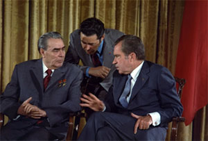 Image of Nixon and Brezhnev seated and one man is leaning down behind them.