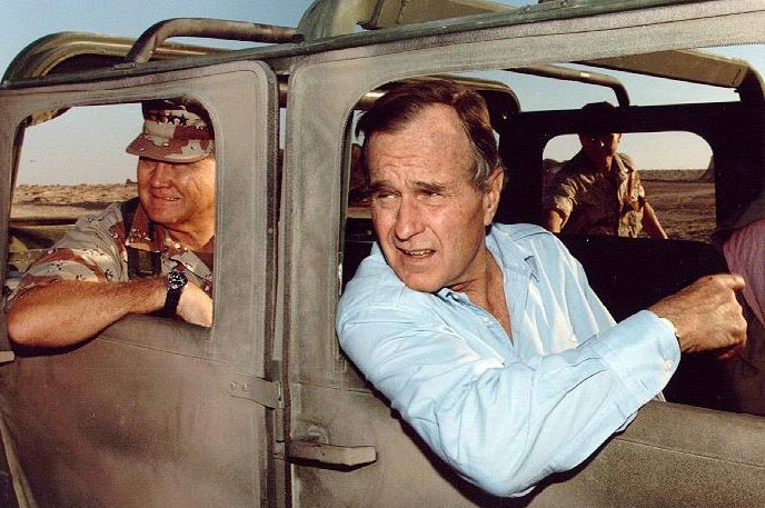 Image of George H.W. Bush riding in a jeep, his arm is hanging out. General Schwarzkopf is riding in the back seat