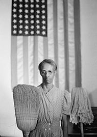 : Image of an African-American woman holding a broom in one hand, and a mop in the other. An American flag is hanging on the wall in the background